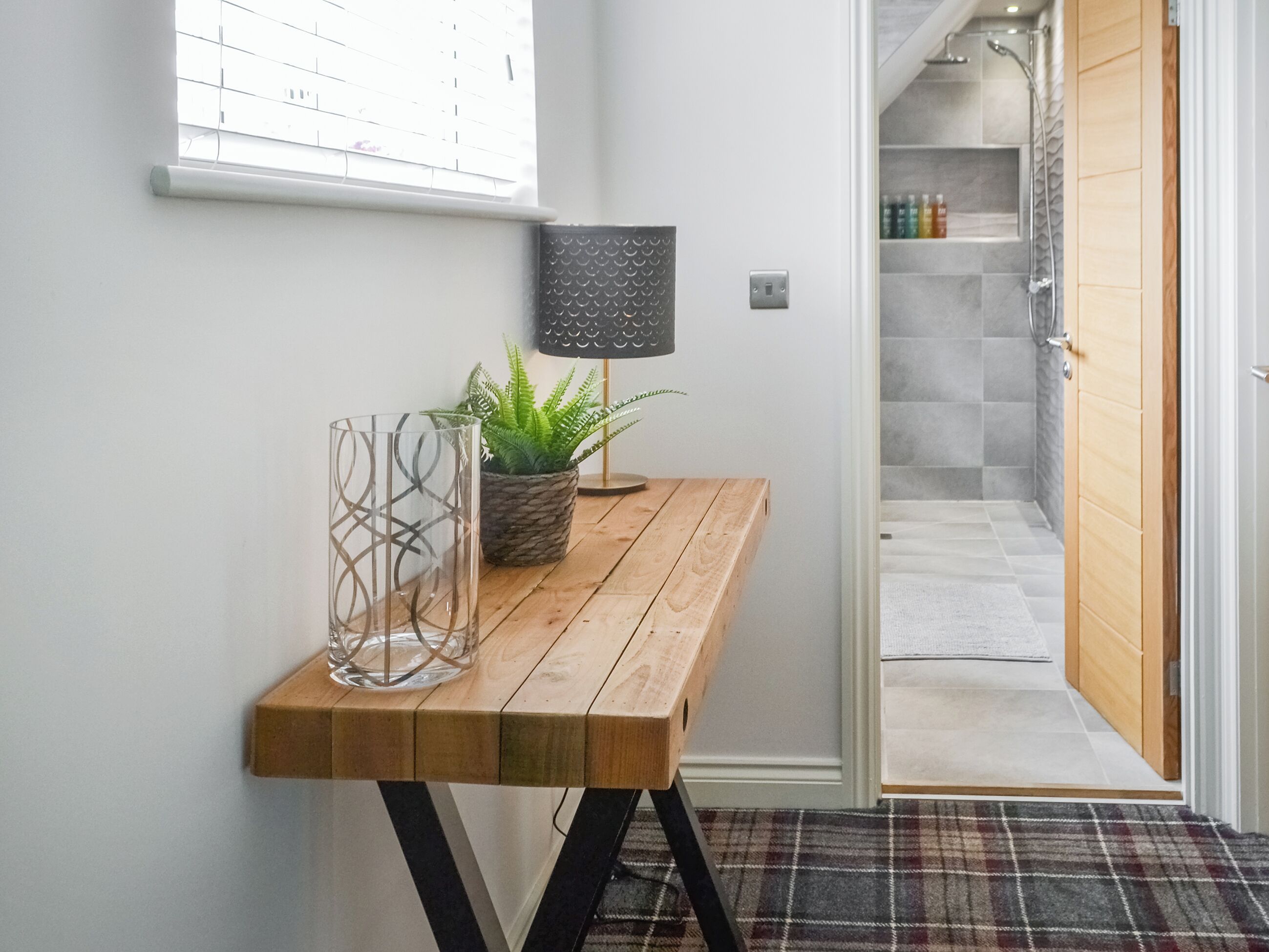 Un couloir avec un banc en bois, une plante en pot, une lampe et une vue sur une salle de bain équipée d’une douche et dont les murs sont recouverts de carreaux.