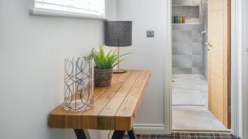 Un couloir avec un banc en bois, une plante en pot, une lampe et une vue sur une salle de bain équipée d’une douche et dont les murs sont recouverts de carreaux.
