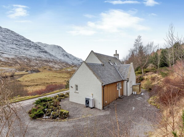A small, single-story house with a sloped roof, a chimney, and a gravel driveway.