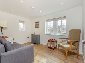 A room with a grey sofa, a wooden chair, a small table with a vase, and a cabinet. There are windows with blinds, a ceiling light, and a picture on the wall.