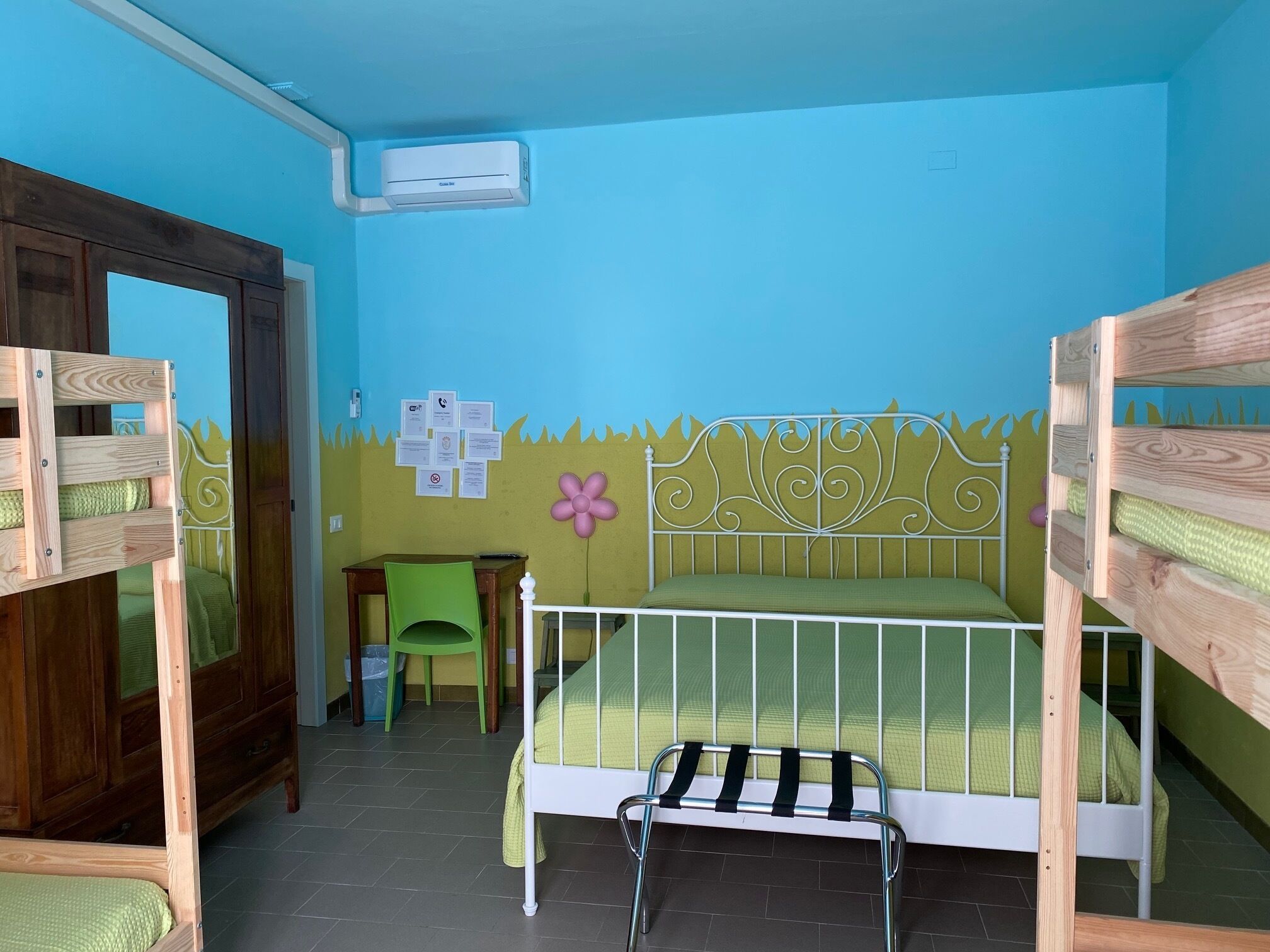 Een kinderkamer met stapelbedden, een houten garderobe, een klein tafeltje en een prullenbak.