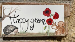 Une enseigne décorative représentant un hérisson, un écureuil et des coquelicots rouges, avec le texte « Happy green » sur un fond de mur en briques.