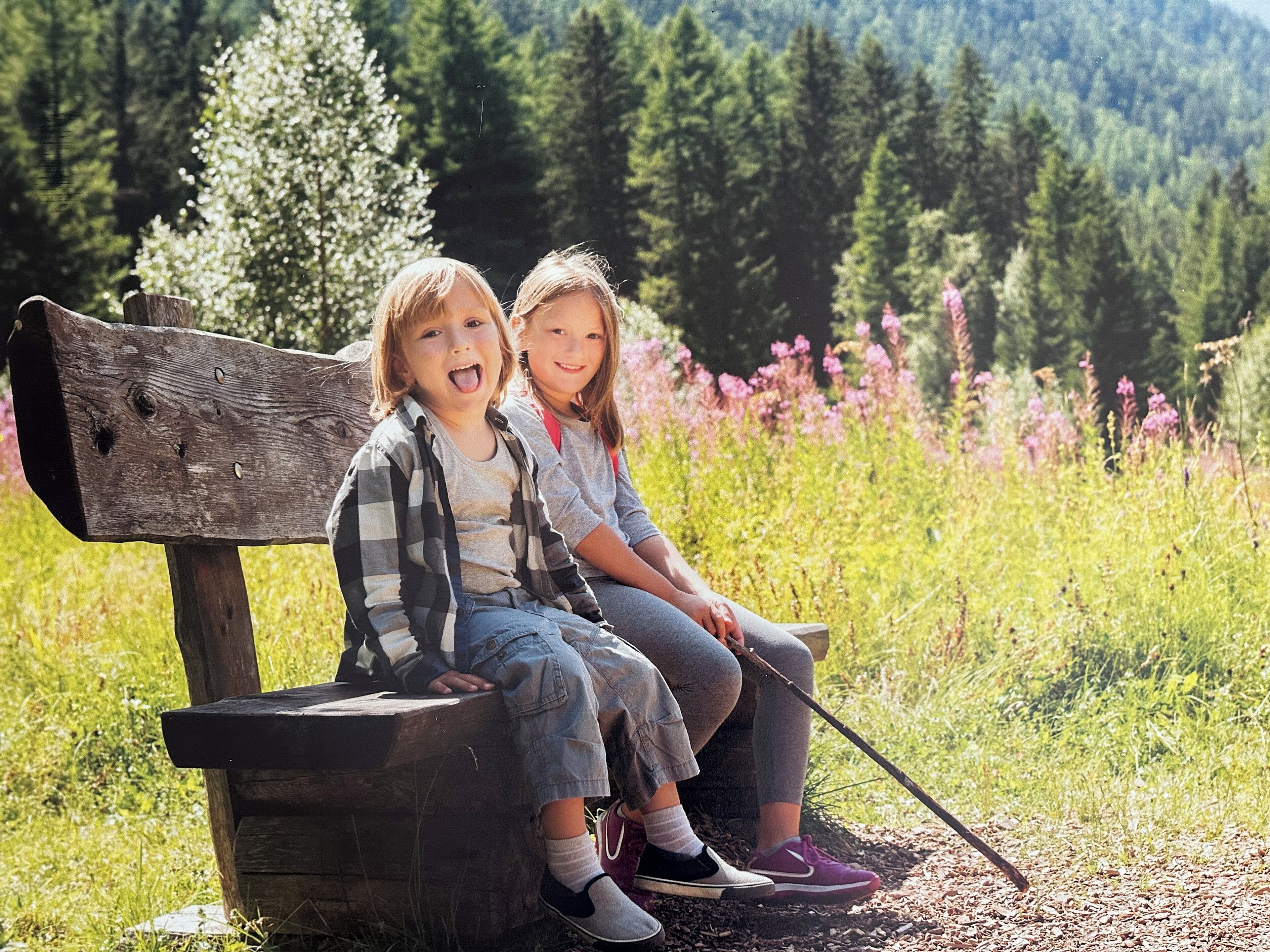 Zwei Kinder sitzen auf einer Holzbank in einer natürlichen Umgebung, im Hintergrund Bäume und Blumen.