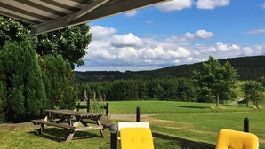 An outdoor patio with a table and two yellow chairs, overlooking a grassy landscape with trees and hills. - Haus Aktiv Winterberg, Garten Apartment, Www.haus-aktiv.de - 2-6 Personen (Winterberg)