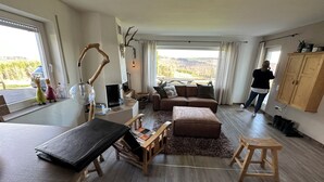 A spacious living room with a large window, a sofa, a coffee table, and a wooden cabinet. - Haus Aktiv Winterberg, Garten Apartment, Www.haus-aktiv.de - 2-6 Personen (Winterberg)