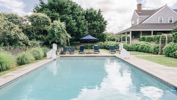 A rectangular swimming pool with a tiled edge, surrounded by a grassy area and a wooden fence. There are lounge chairs and a blue umbrella in the background. - Ultimate relaxation spot with steam room, close to the beach. Family-friendly (Nantucket)