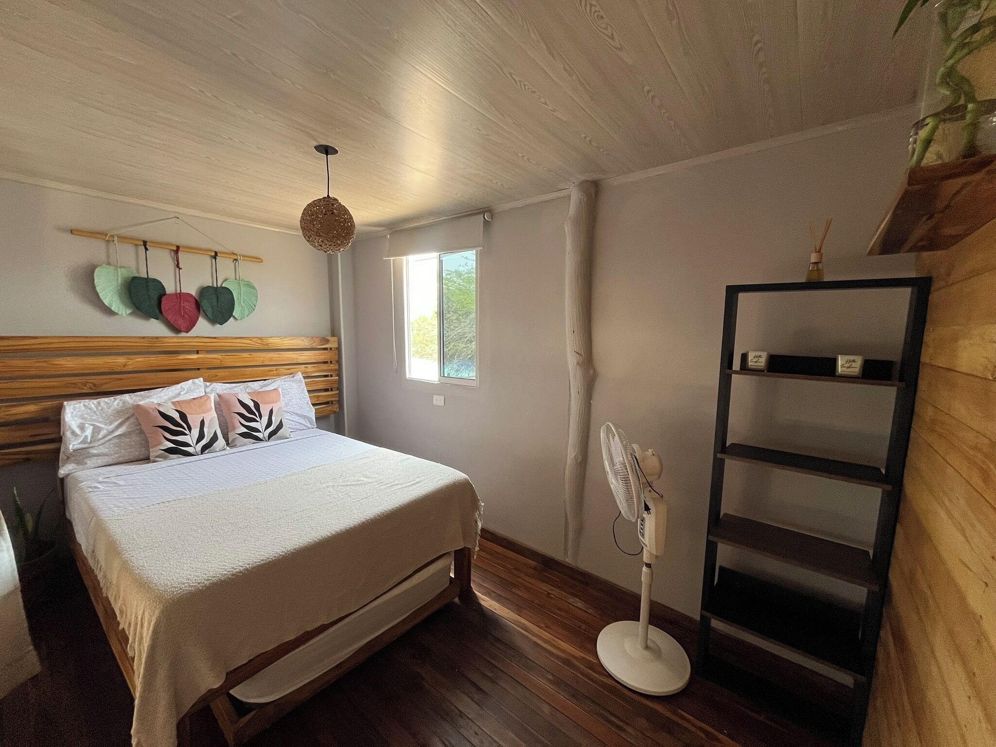 A bedroom with a wooden bed, a bedside fan, a window, and a shelf with decorative items.
