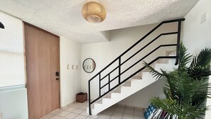 Una escalera con barandilla negra que sube a un piso superior, una puerta de madera, un espejo y una planta en maceta a la derecha.