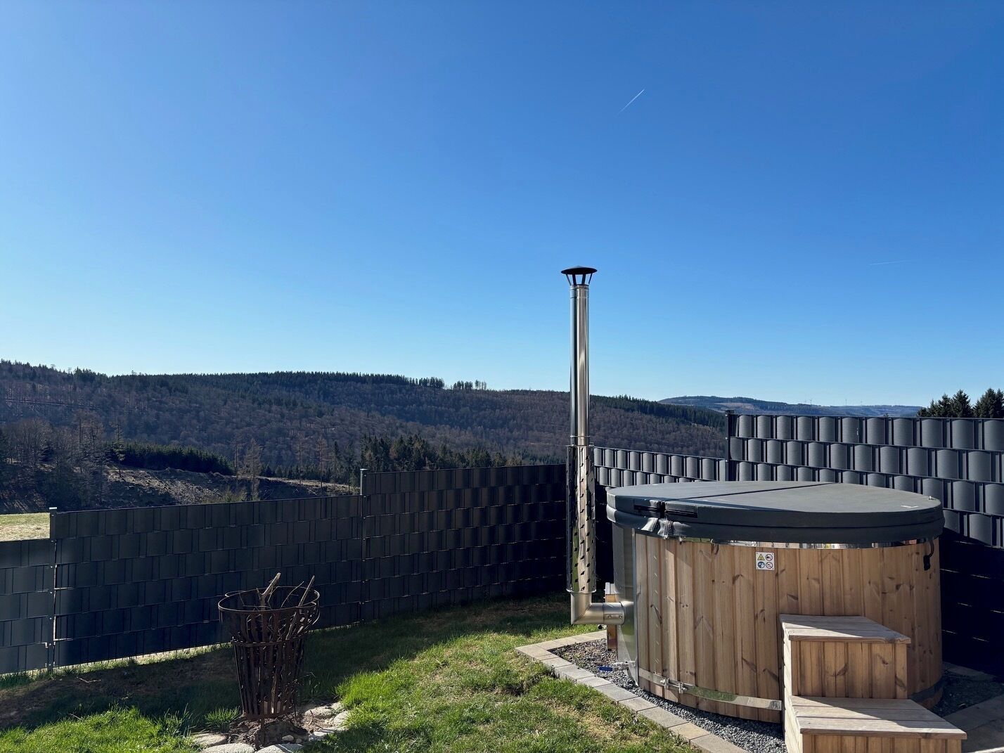 Ett bubbelbad med en trätrall och en metallvärmare, omgivet av ett metallstaket, med utsikt över en skogbeklädd kulle i bakgrunden.