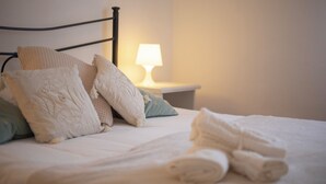 Een net opgemaakt bed met kussens en een opgerolde handdoek, een bedlamp en een nachtkastje.