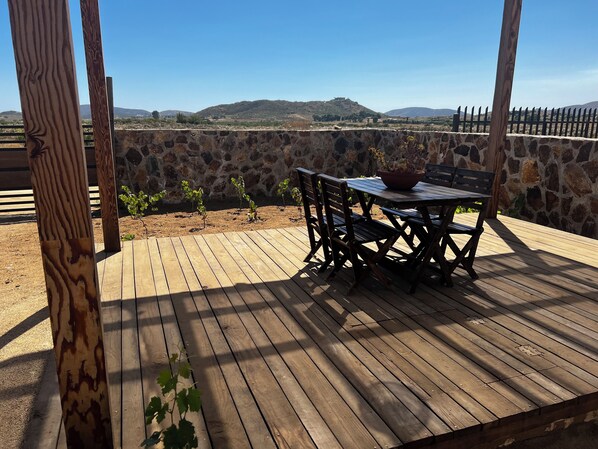A wooden deck with a table and chairs, surrounded by stone walls and a view of distant hills.