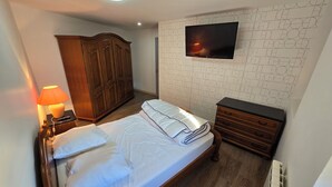 Un lit bien fait, avec des oreillers, une armoire en bois, une table de chevet avec une lampe, un téléviseur fixé au mur et un radiateur.