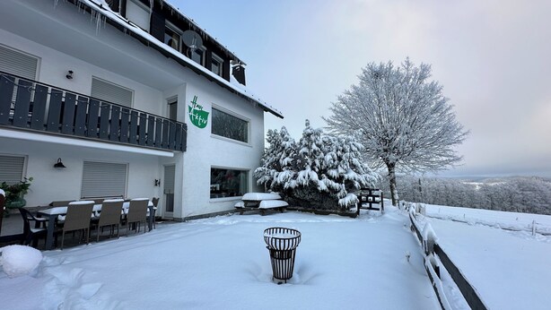 Un espace extérieur enneigé avec un bâtiment, des sièges en plein air et un arbre recouvert de neige.