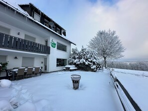 A snow-covered outdoor area with a building, outdoor seating, and a snow-covered tree. - 3-Bedroom Apartment in Winterberg (Winterberg)