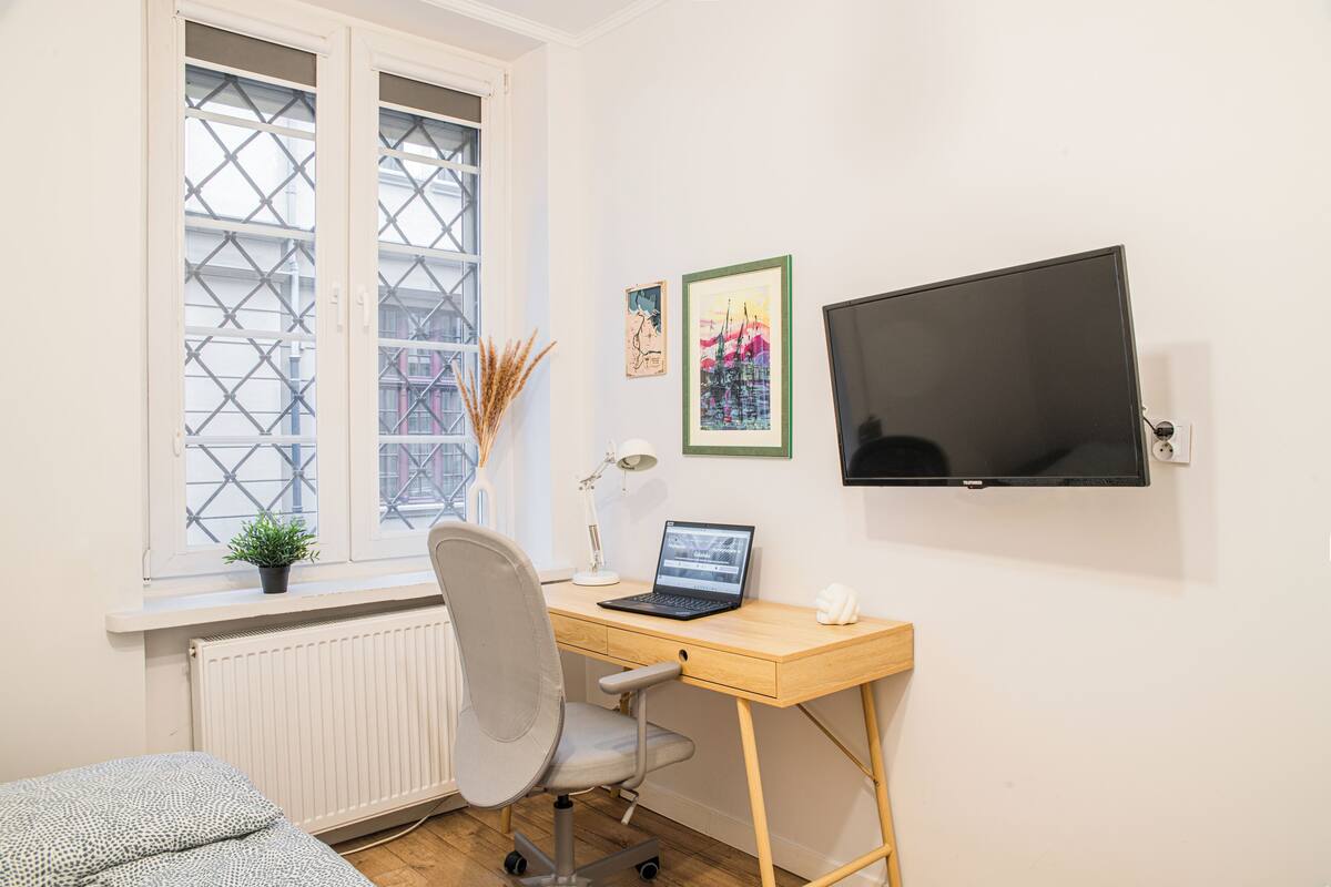 A room with a wooden desk, a laptop, a chair, a radiator, a window with a view, and a TV on the wall.