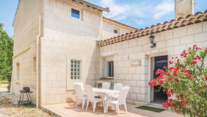 Un edificio de piedra con techo de tejas, un patio con mesa y sillas, y un pequeño jardín con flores rojas.