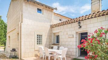 Un edificio in pietra con tetto in tegole, un patio con tavolo e sedie, e un piccolo giardino con fiori rossi.