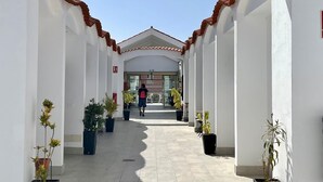 Un couloir aux murs blancs, au sol carrelé et au toit recouvert de tuiles. Des plantes en pot sont disposées le long des murs, et une personne marche en direction de l’entrée d’un bâtiment.