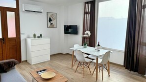 Un salon moderne comprenant un canapé, une table à manger avec des chaises, une table basse, un ventilateur de plafond, un appareil de climatisation, un téléviseur et une grande fenêtre avec des rideaux.