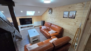 Ein gemütliches Wohnzimmer mit einem braunen Ledersofa, einem weißen Couchtisch, einem Fernseher auf einem Schrank, holzverkleideten Wänden und einer Uhr.