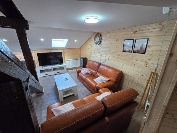 A cozy living room with a brown leather sofa, a white coffee table, a TV on a stand, wooden panel walls, and a clock.