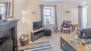 A living room with a fireplace, a television, a sofa, a coffee table, and a bookshelf. - Discover coastal tranquility in Millhouse Cottage on Budle Bay. (Bamburgh)