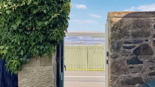 A stone wall with a green door leading to a beach view. - Discover coastal tranquility in Millhouse Cottage on Budle Bay. (Bamburgh)