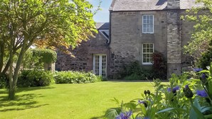 A stone building with a well-maintained lawn and flowering plants. - Discover coastal tranquility in Millhouse Cottage on Budle Bay. (Bamburgh)