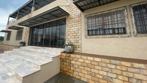 A modern two-story house with a stone facade, large windows with decorative iron grilles, and a covered outdoor area with steps leading to a patio.