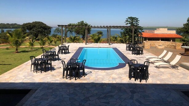 A swimming pool with surrounding chairs and tables, a pergola, and a view of the sea.
