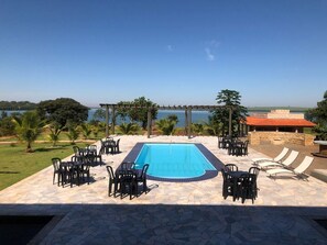 A swimming pool with surrounding chairs and tables, a pergola, and a view of the sea.