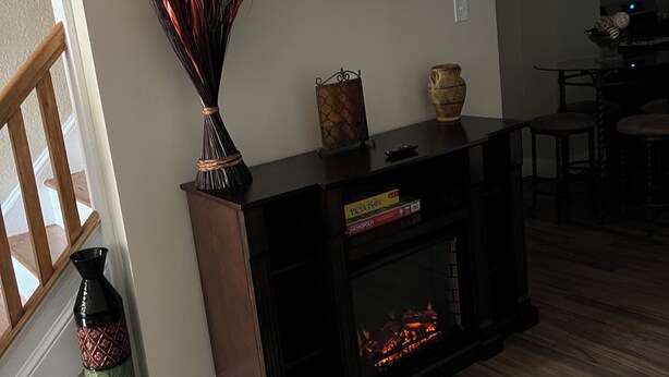 Una sala de estar con chimenea, un televisor de pantalla plana montado en la pared, una escalera de madera y un comedor al fondo.