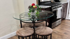 Una mesa redonda de vidrio con cuatro taburetes acolchados, un microondas de acero inoxidable en la parte superior, una estufa a gas y una cafetera sobre la encimera.