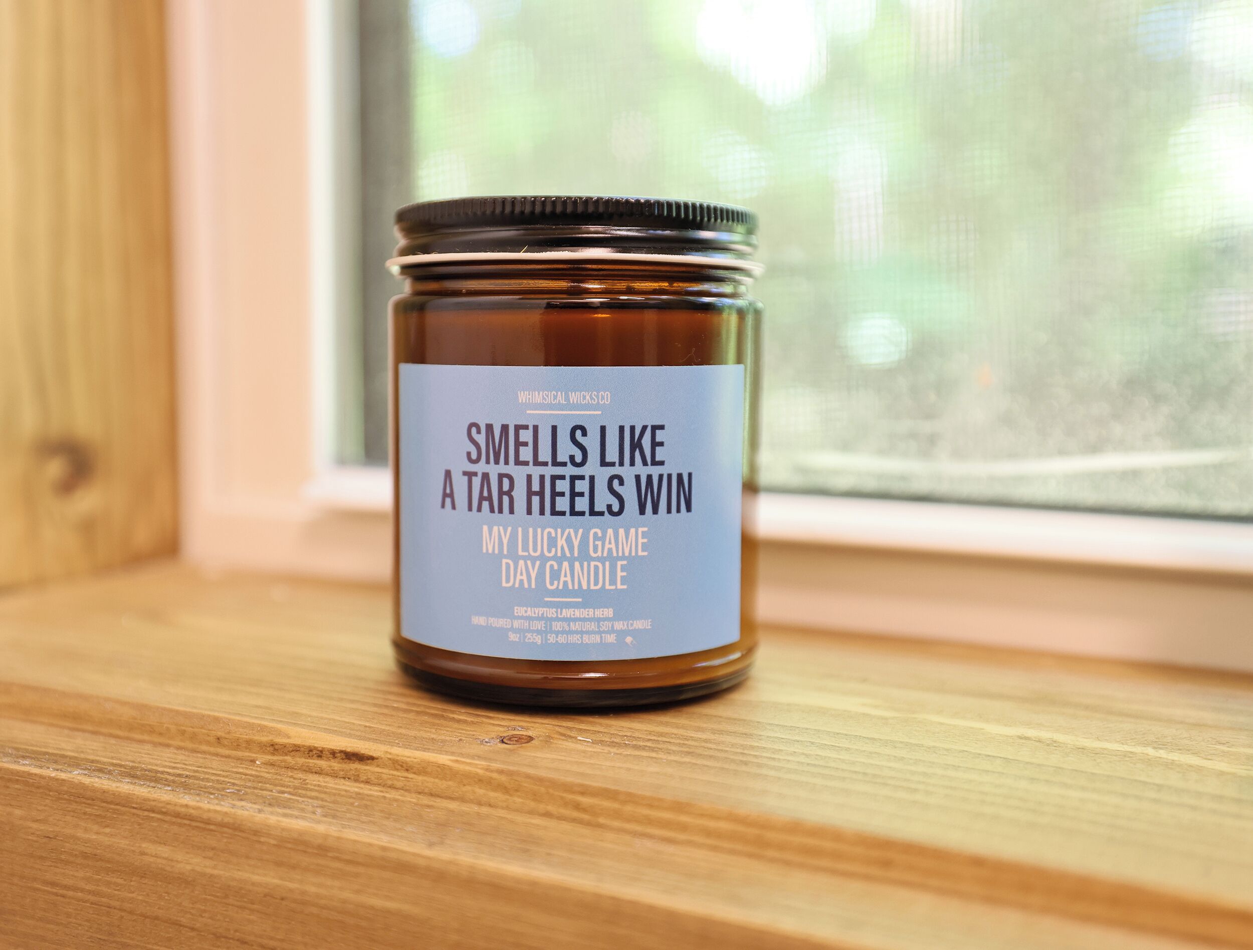 A jar of "Smells Like a Tar Heels Win" day candle on a wooden surface near a window.