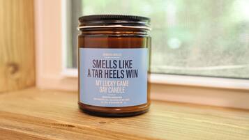 A jar of "Smells Like a Tar Heels Win" day candle on a wooden surface near a window.