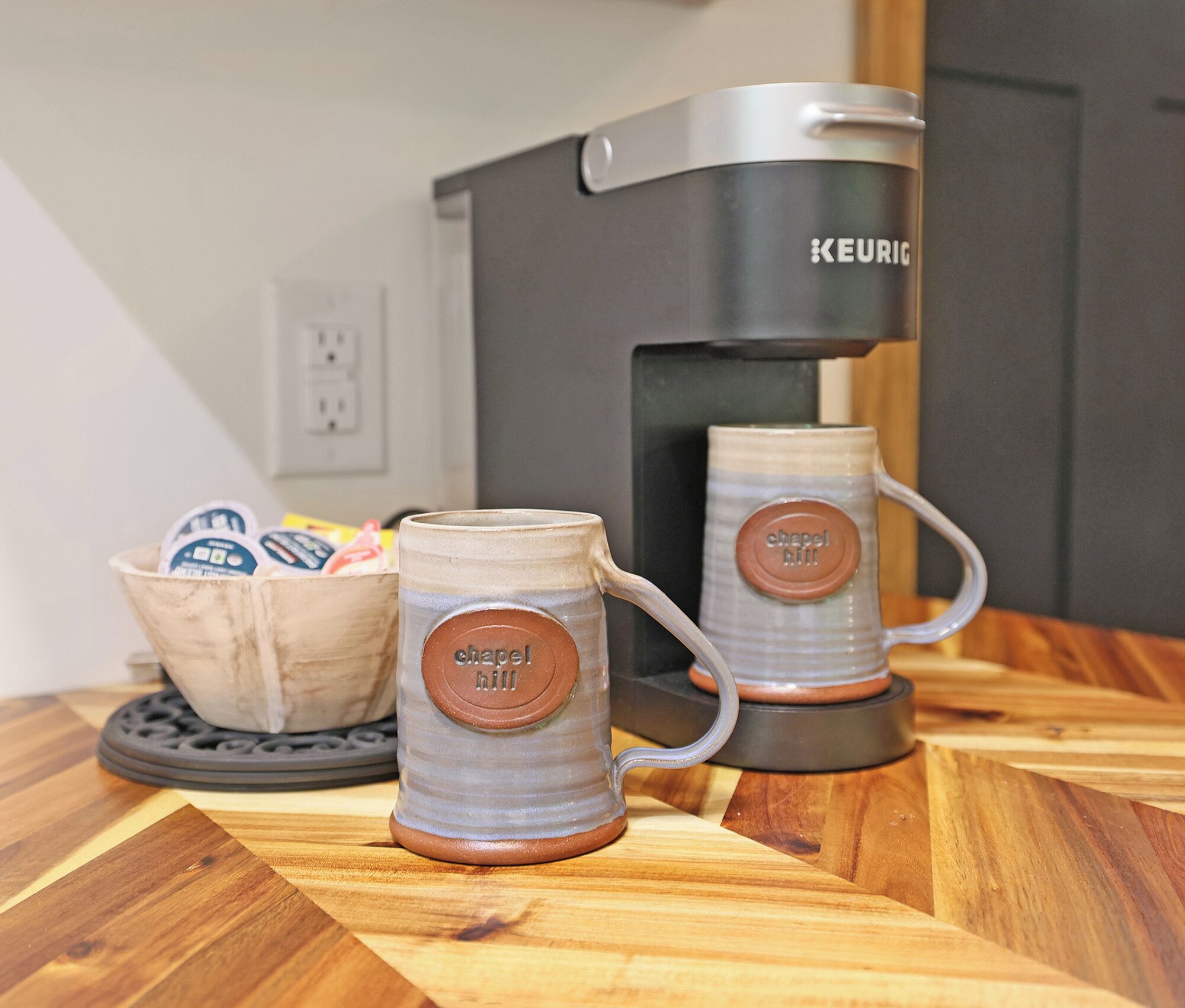 Two ceramic mugs with "chapel hill" branding on a wooden table, a Keurig coffee maker, and a basket with coffee pods.