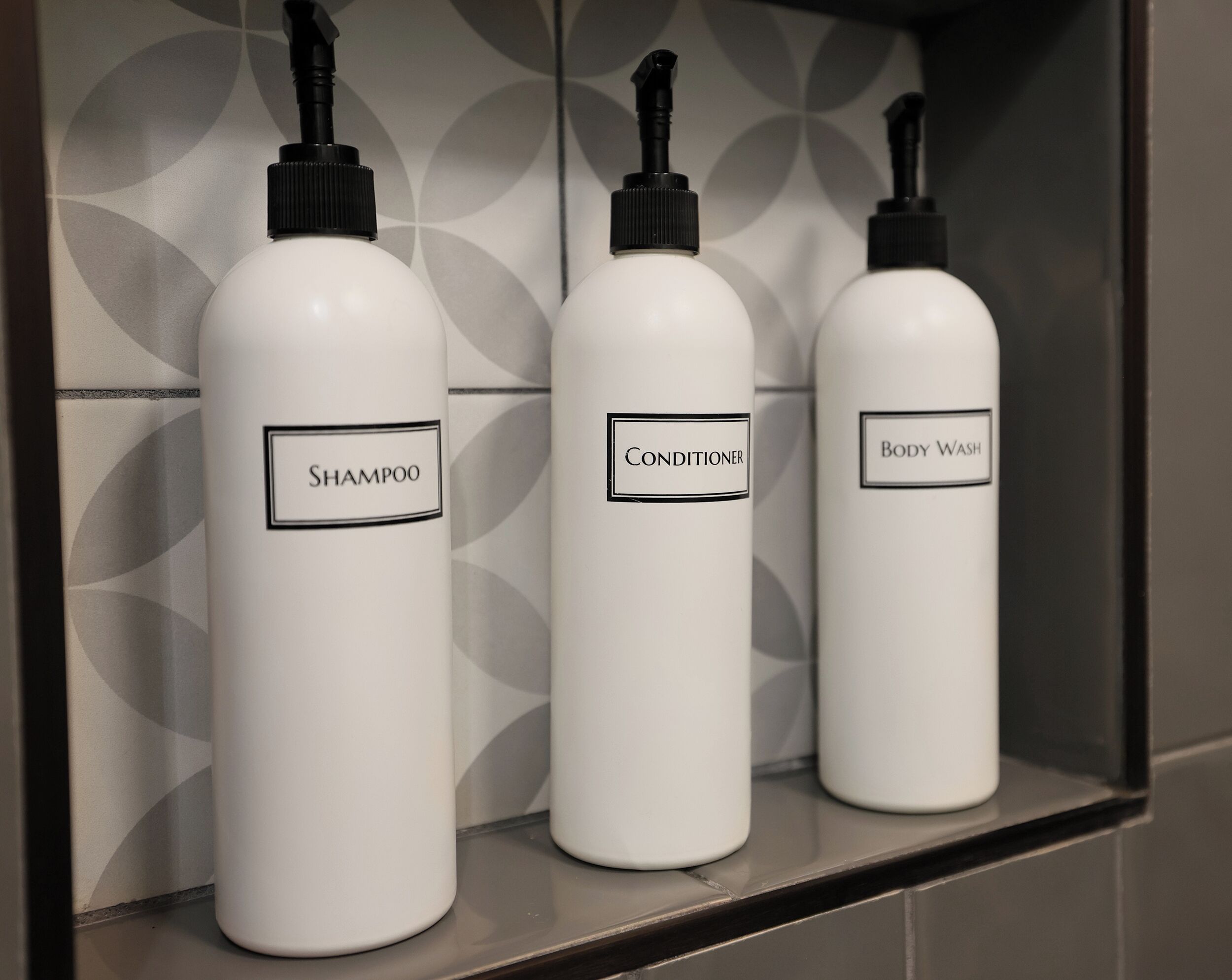 Tres botellas blancas etiquetadas como champú, acondicionador y gel de baño sobre una estantería.