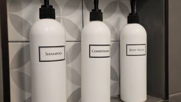 Tres botellas blancas etiquetadas como champú, acondicionador y gel de baño sobre una estantería.