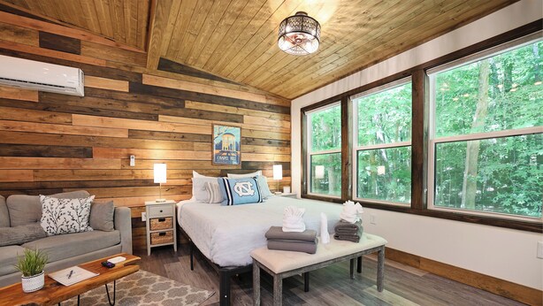 A cozy room with a bed, a sofa, a bench, and a coffee table. There are large windows with a view of trees.