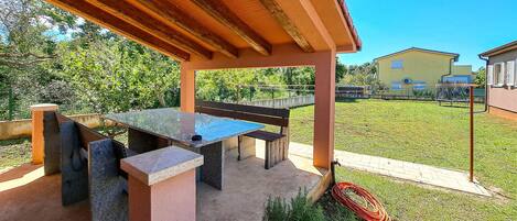 Eine Terrasse im Freien mit Tisch und Bänken, umgeben von einer Rasenfläche und einem Gebäude im Hintergrund.