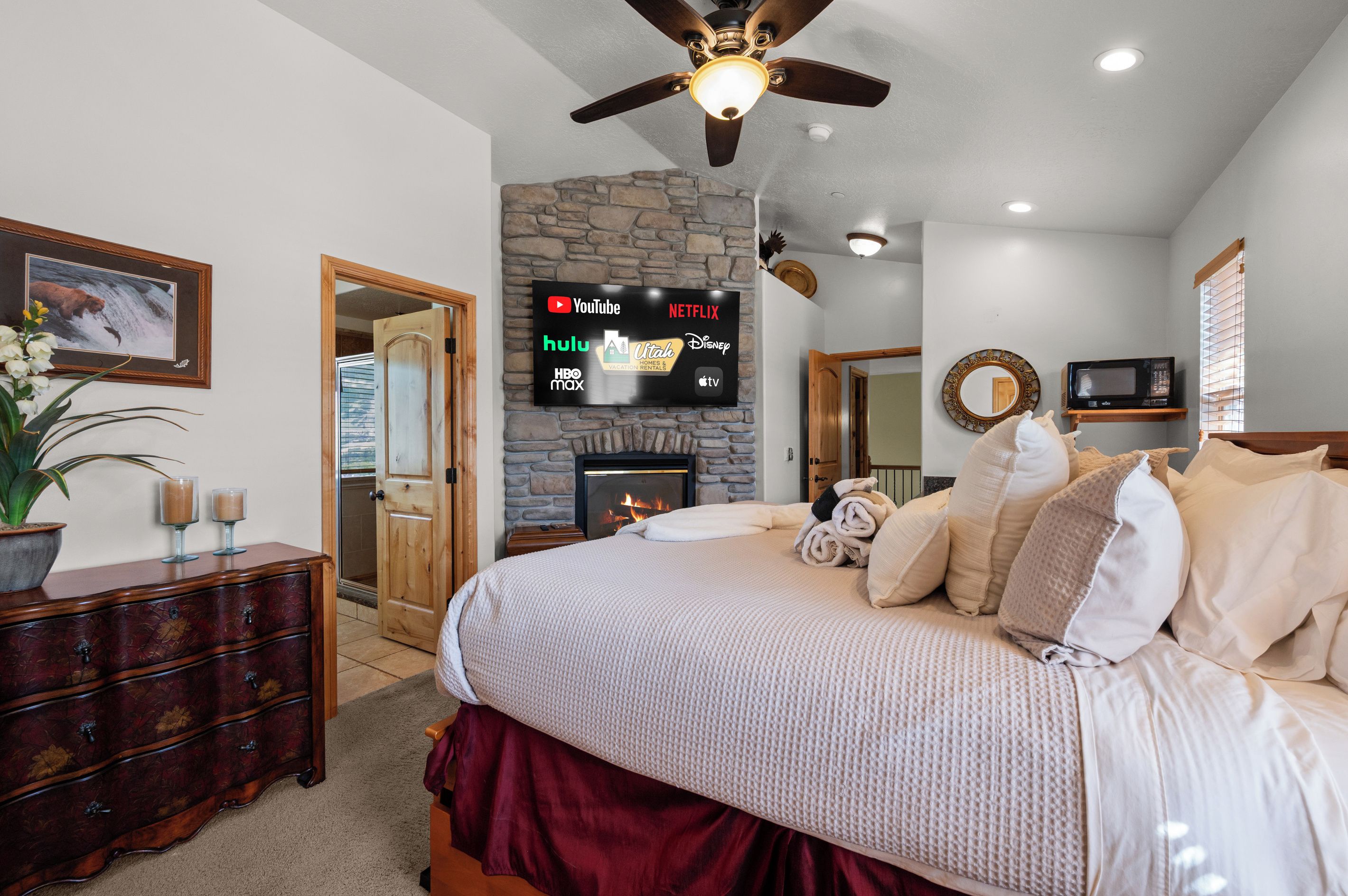 A bedroom with a stone fireplace, a bed with multiple pillows, a dresser, and a television mounted on the wall displaying various streaming services.