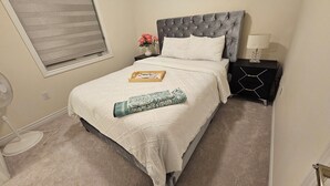 Ein ordentlich bezogenes Bett mit weißer Daunendecke, einem grauen, gepolsterten Kopfteil und einem Nachttisch mit Lampe.
