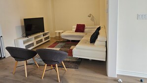 Ein modernes Wohnzimmer mit Fernseher, einem weißen Sofa, zwei schwarzen Sesseln und einem kleinen Beistelltisch.
