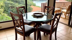 A round glass table with four wooden chairs, a wooden deck with a railing, and a scenic view of a river and forest through large windows. - Beautiful Ocean View, steps to the Beach, 3 Pools, Los Almendros #31 (Playa Ocotal)