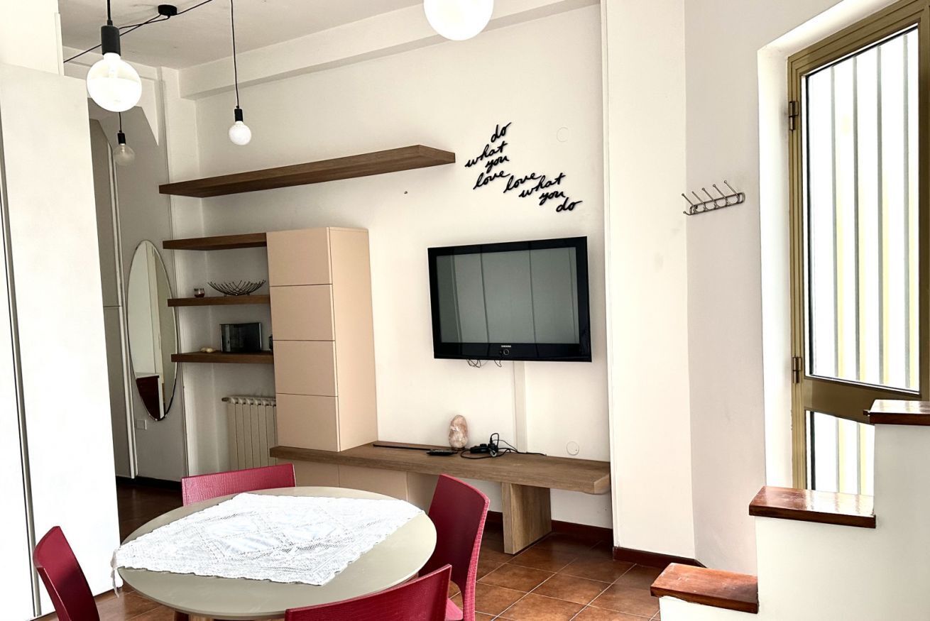 Uma sala de estar moderna com uma mesa redonda, cadeiras vermelhas, uma TV de tela plana e uma prateleira fixada na parede.