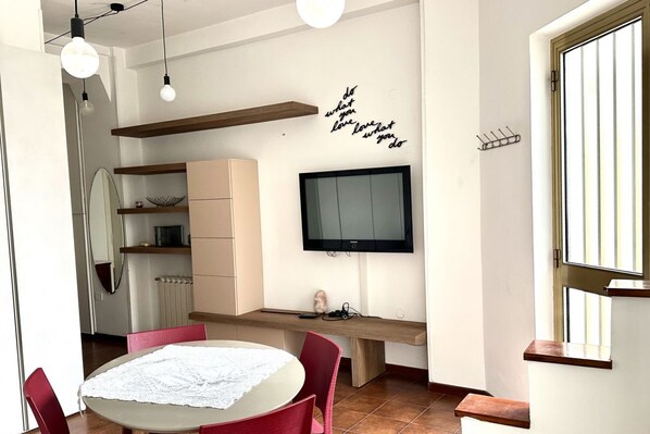 A modern living room with a round table, red chairs, a flat-screen TV, and a wall-mounted shelf.