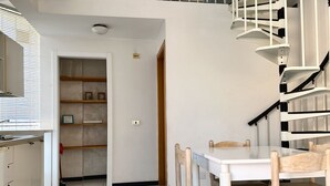 Et køkken med hvide skabe, et spisebord med træstole, en trappe med hvide gelændere og en dør der fører til en gang.