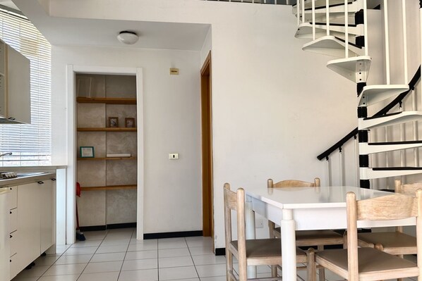 Une cuisine avec des meubles blancs, une table à manger entourée de chaises en bois, un escalier avec des rampes blanches et une porte donnant sur un couloir.