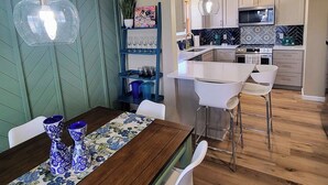 Eine moderne Küche mit weißen Schränken, einer Mikrowelle und einer Kaffeemaschine. Ein Essbereich mit einem Holztisch und einer blau gemusterten Tischdecke.