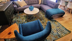 Ein Wohnzimmer mit einem Ecksofa, einem runden Couchtisch, einem blauen Sessel, einem gemusterten Teppich und einem Bild an der Wand.